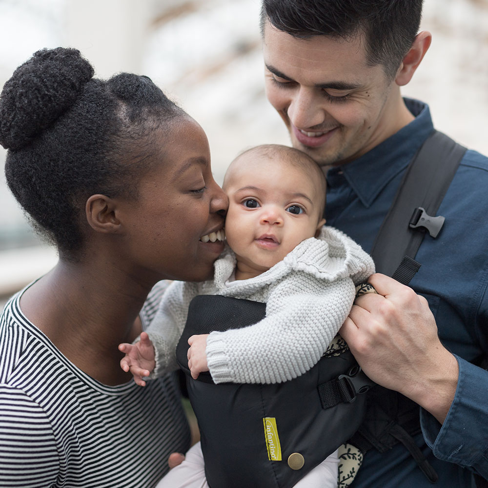 A black woman kisses a baby behind held in a carrier by her partner