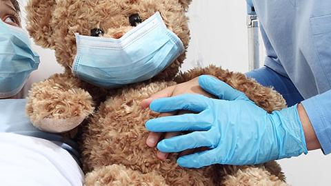 Woman in mask gives teddy bear wearing mask to child in hospital bed wearing mask