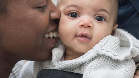 A black woman kisses a baby behind held in a carrier by her partner